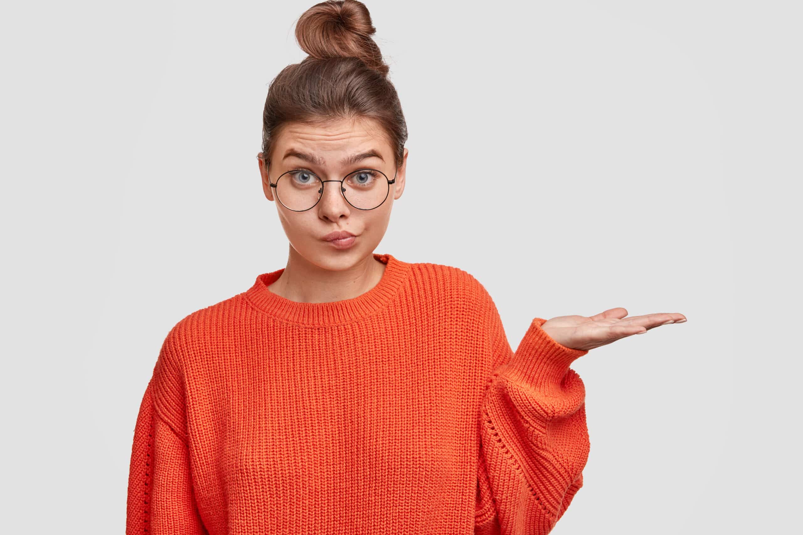 Une jolie mannequin perplexe et hésitante, les cheveux attachés en chignon, vêtue d'un pull oversize, lève la main près de son épaule, pointe du doigt un espace vierge destiné à votre contenu publicitaire et esquisse un sourire étonné